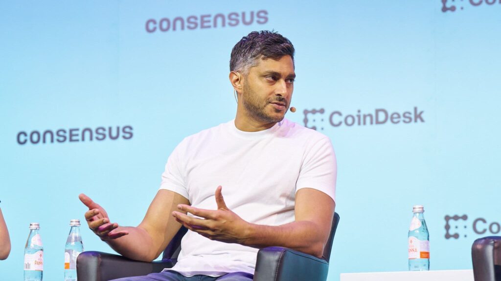 no_translation_1403_ffhaj7cer7v0iim75vzkfy4c_no_translation_translation - Crypto and Coin Man in a white T-shirt speaks on a panel with hands gesturing, blue backdrop with 'CONSENSUS' branding and water bottles on the table