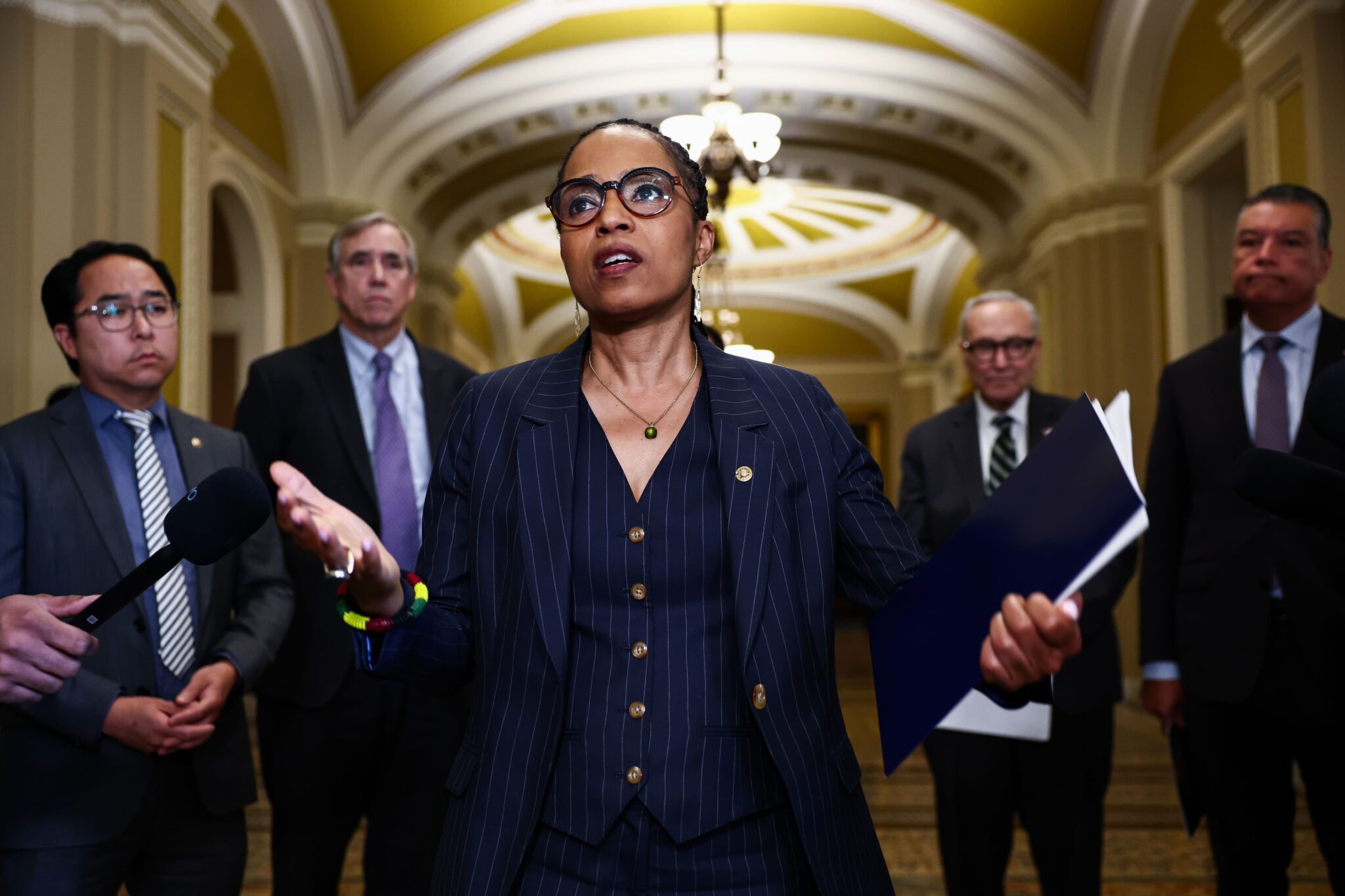 Central woman in a navy pinstripe suit speaks to reporters in a grand hallway, microphones nearby, colleagues around her.
