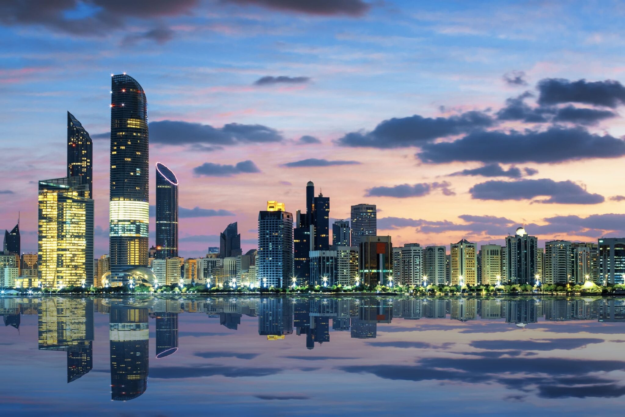 City skyline at dusk with illuminated skyscrapers and a calm water reflection.