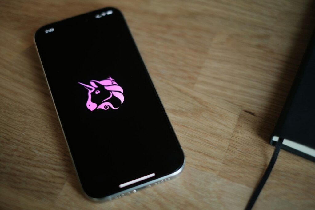 no_translation_2228_ffhaj7cer7v0iim75vzkfy4c_no_translation_translation - Crypto and Coin Smartphone lying on a wooden desk with a pink unicorn logo displayed on its black screen, close-up view.