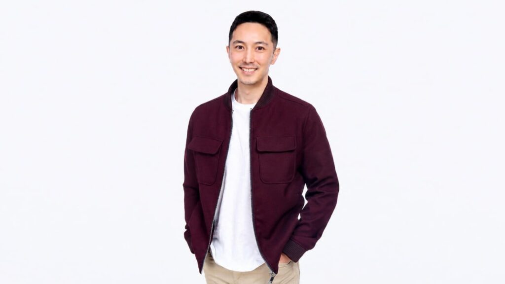 Smiling man in a burgundy jacket over a white T-shirt, beige pants, standing against a plain white background.