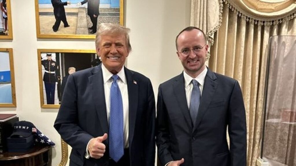 Two men in suits posing indoors; one gives a thumbs-up, with framed pictures and gold curtains in the background.