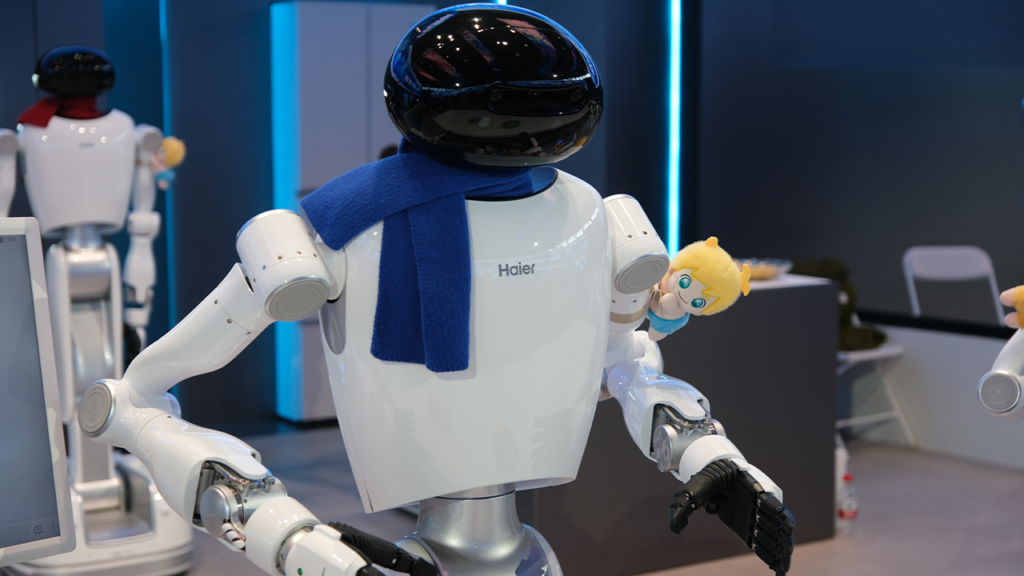 White Haier robot wearing a blue scarf, holding a small plush doll with blond hair.
