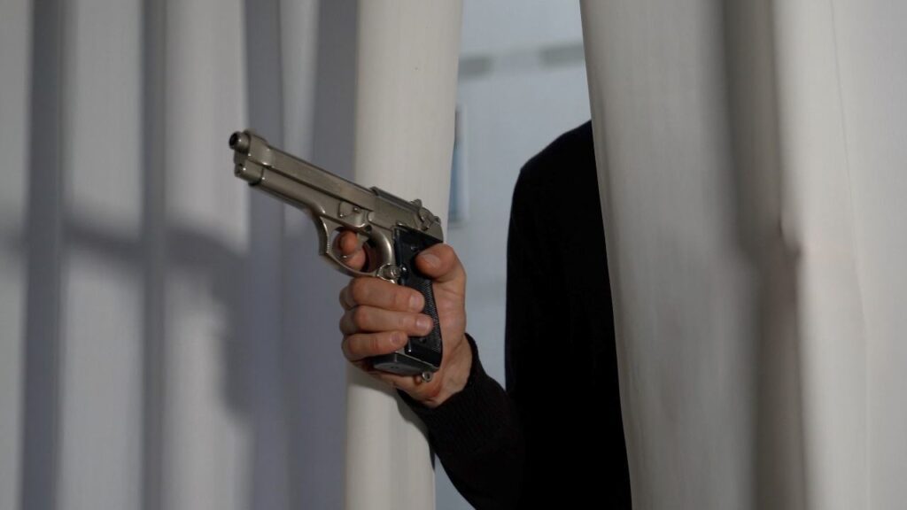 A hand gripping a silver semi-automatic pistol emerging from between white curtains.