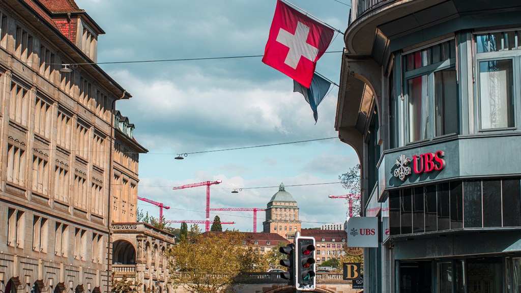 City street with a Swiss flag overhead, UBS storefront on the right, and red construction cranes in the distance under a cloudy sky.